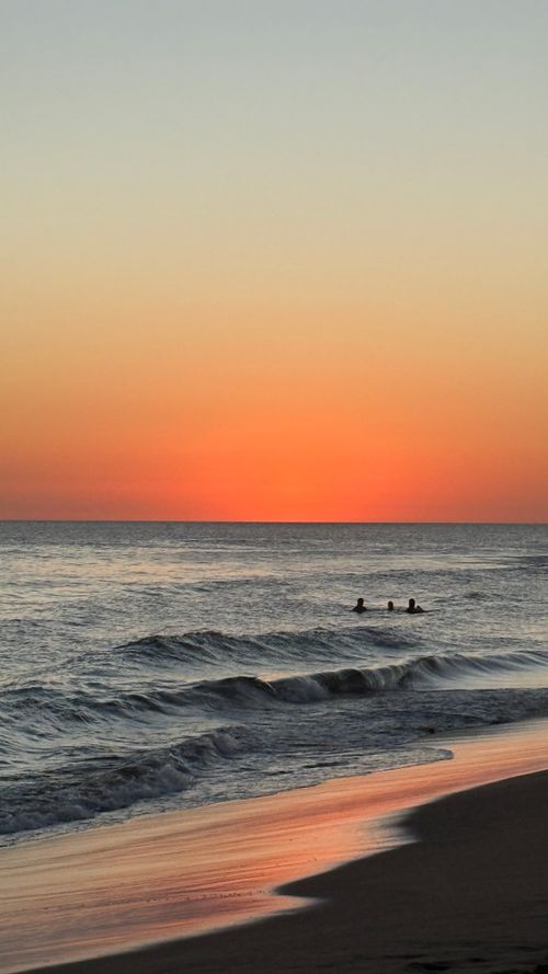 Free A tranquil beach at sunset with three silhouettes swimming in the ocean's gentle waves. Stock Photo