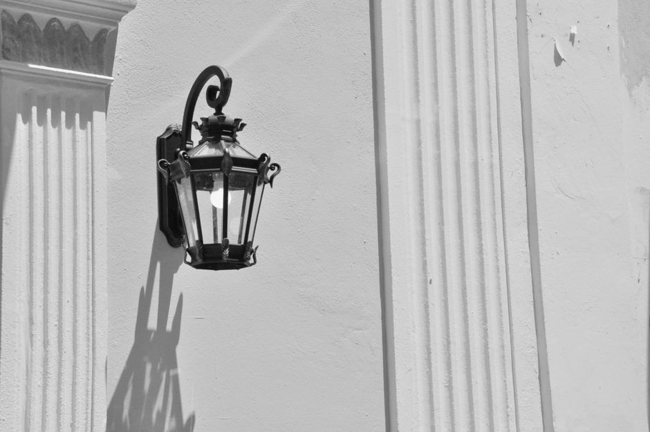 A black and white image of a vintage wall lantern casting distinct shadows on a textured wall.