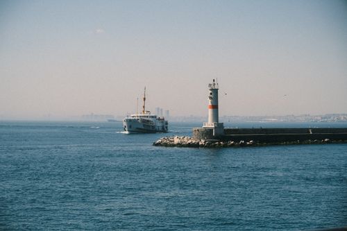 Free Lighthouse by the sea with a ship sailing nearby, offering a tranquil maritime scene. Stock Photo