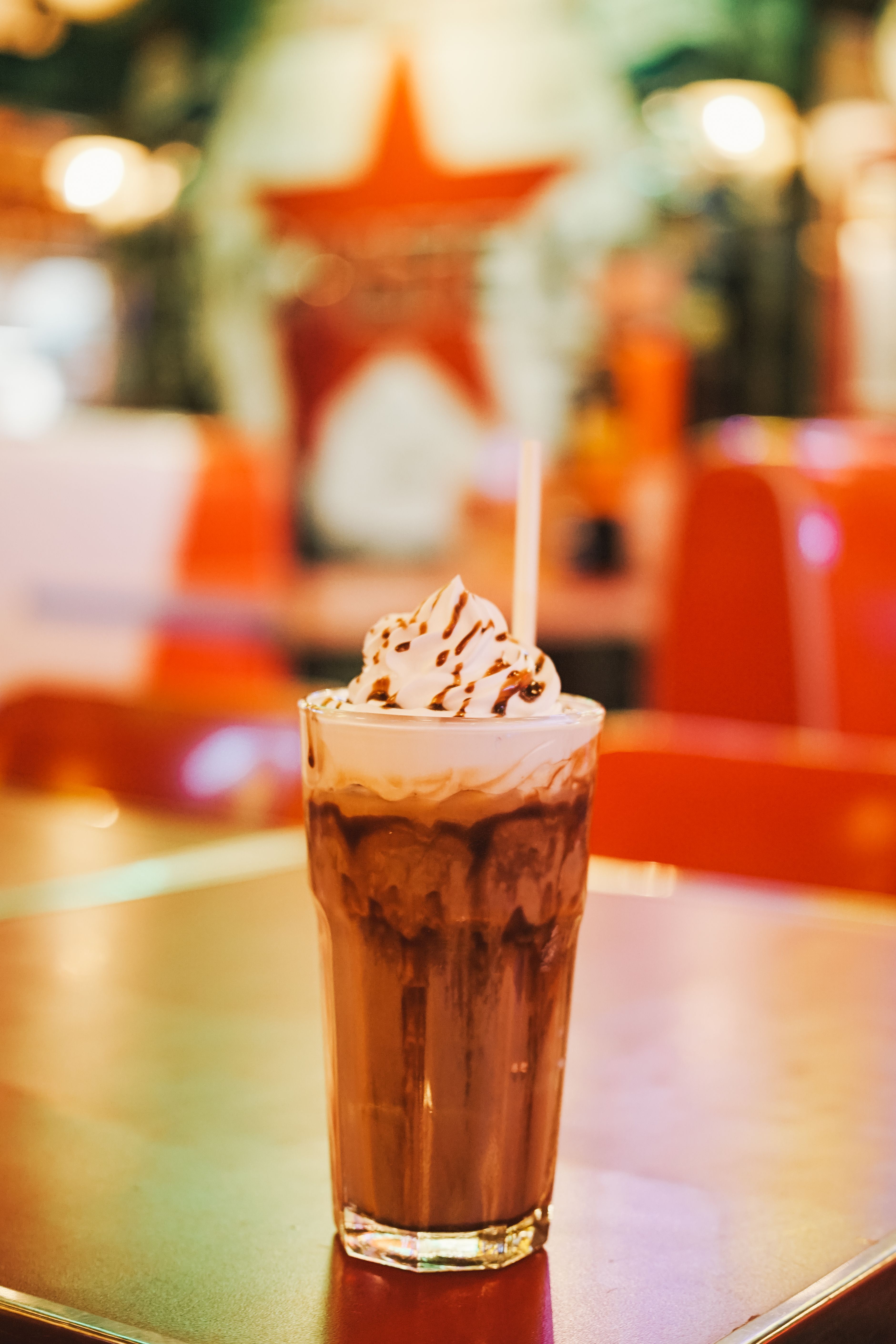 Milkshake au chocolat coiffé de crème fouettée et de sauce, paille blanche, banquettes rouges floues en fond