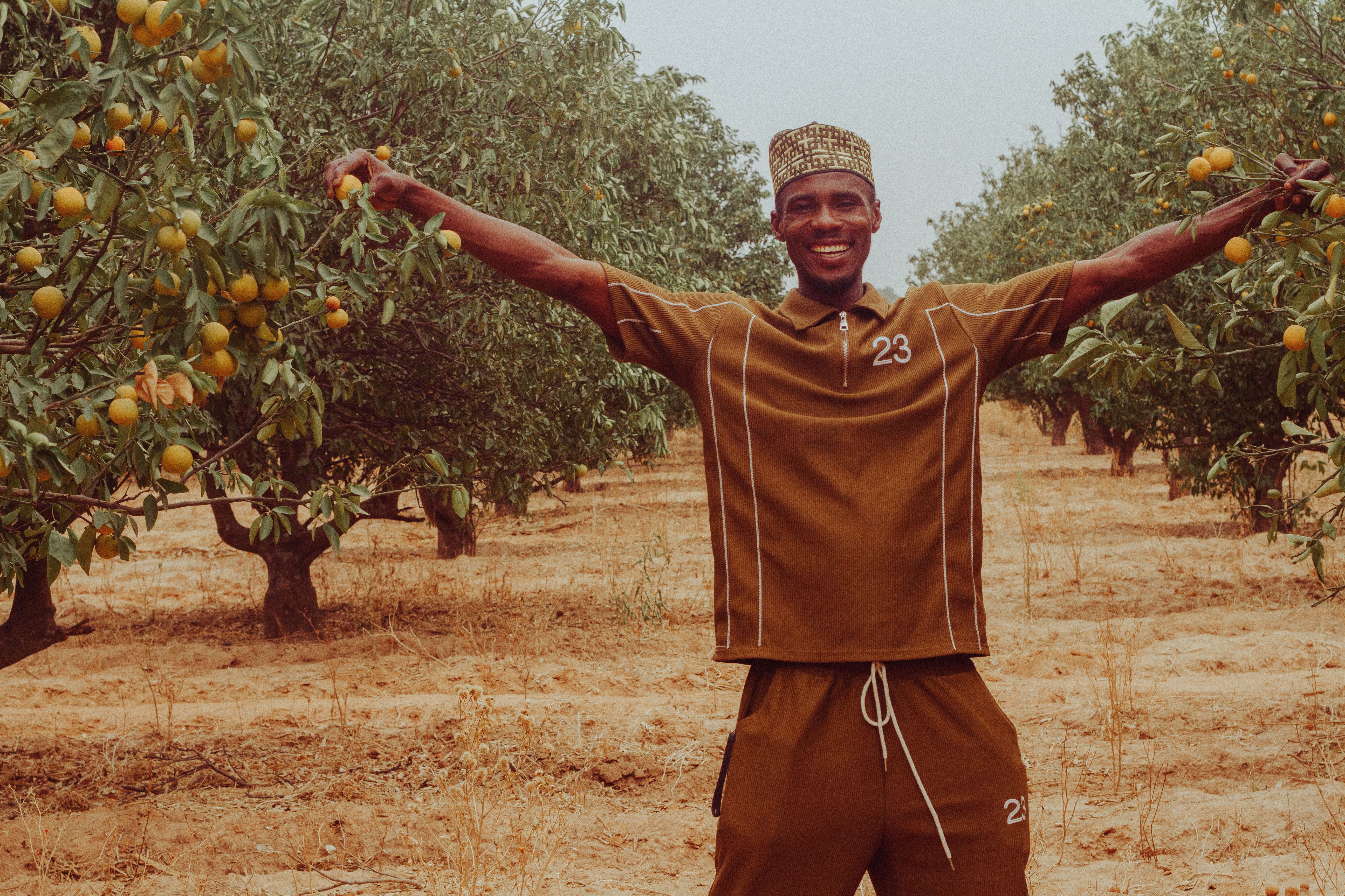 A smiling farmer stretches arms wide amidst lush orange trees in Zaria City, Nigeria.