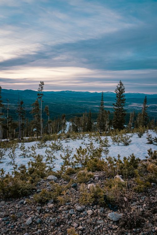 Free A serene view of a mountainous landscape with trees and snow under a colorful twilight sky. Stock Photo