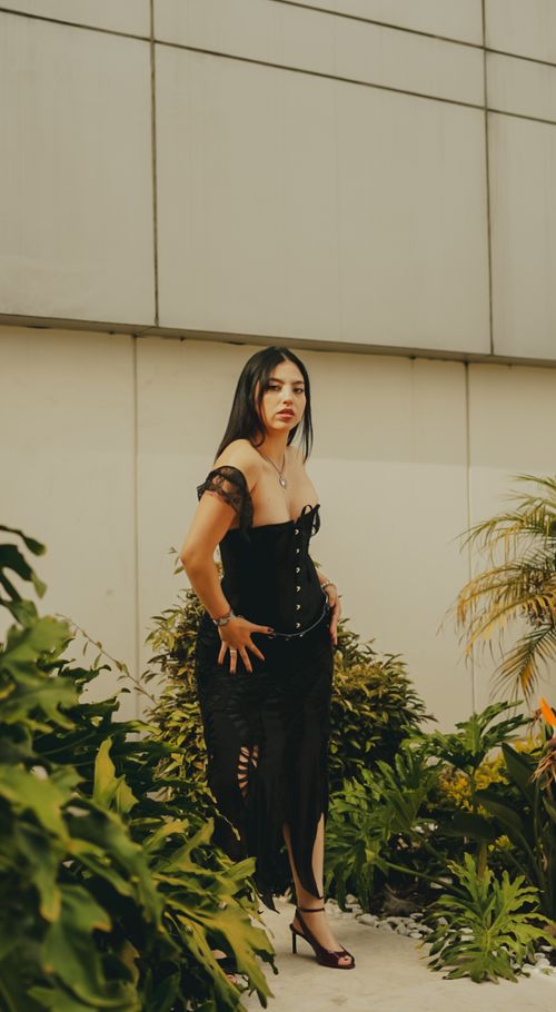 Free Stylish woman in an elegant black dress poses amidst lush greenery and concrete walls. Stock Photo