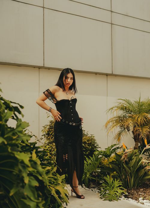Free Stylish woman in black corset dress striking a pose in a modern outdoor setting with lush greenery. Stock Photo