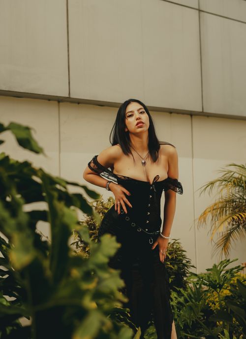 Free Confident woman in a black dress poses outdoors surrounded by lush green plants. Stock Photo