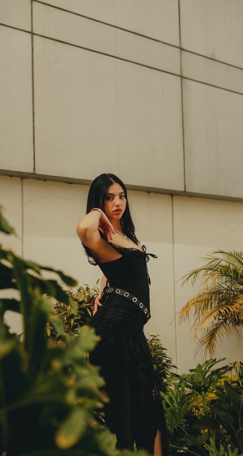 Free Fashionable woman in a black dress posing near lush greenery in an urban environment. Stock Photo