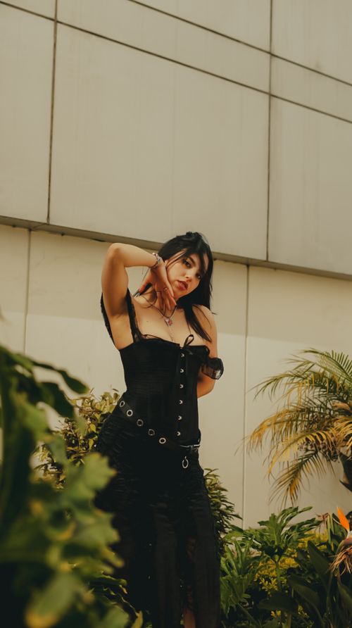 Free Stylish woman posing in a black corset against a building backdrop, surrounded by greenery. Stock Photo