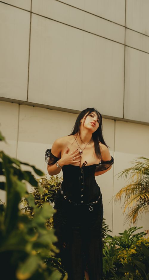 Free A fashionable woman poses in a black corset surrounded by lush greenery against a modern building backdrop. Stock Photo