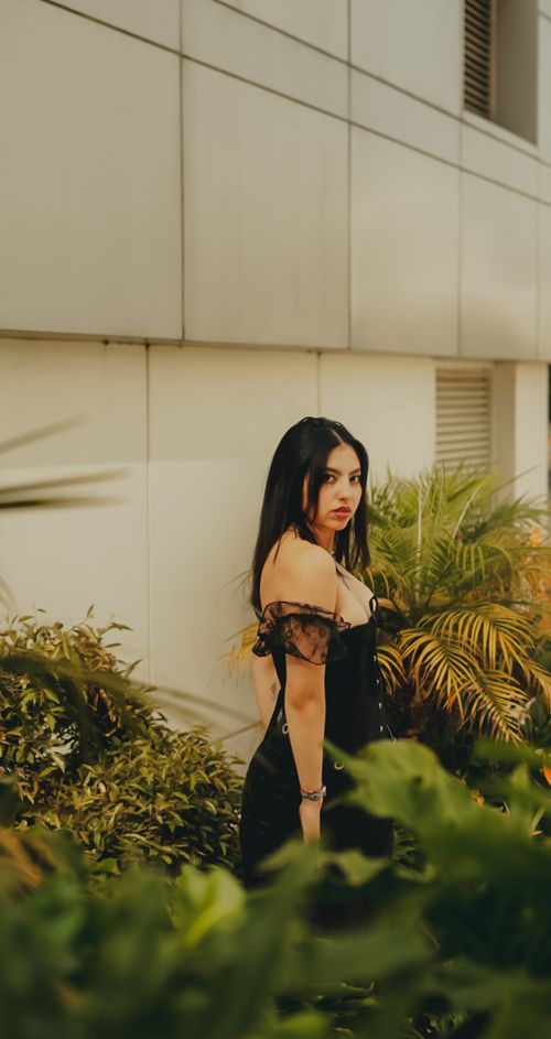 Free Elegant woman in black dress standing amidst lush green plants against modern building backdrop. Stock Photo
