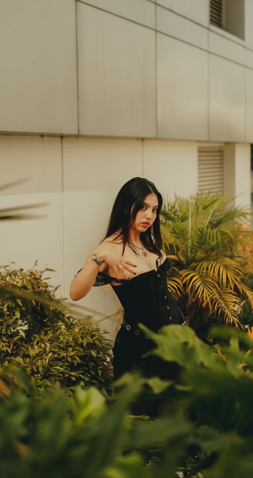 Free A fashionable woman poses confidently in a black dress surrounded by lush urban greenery. Stock Photo