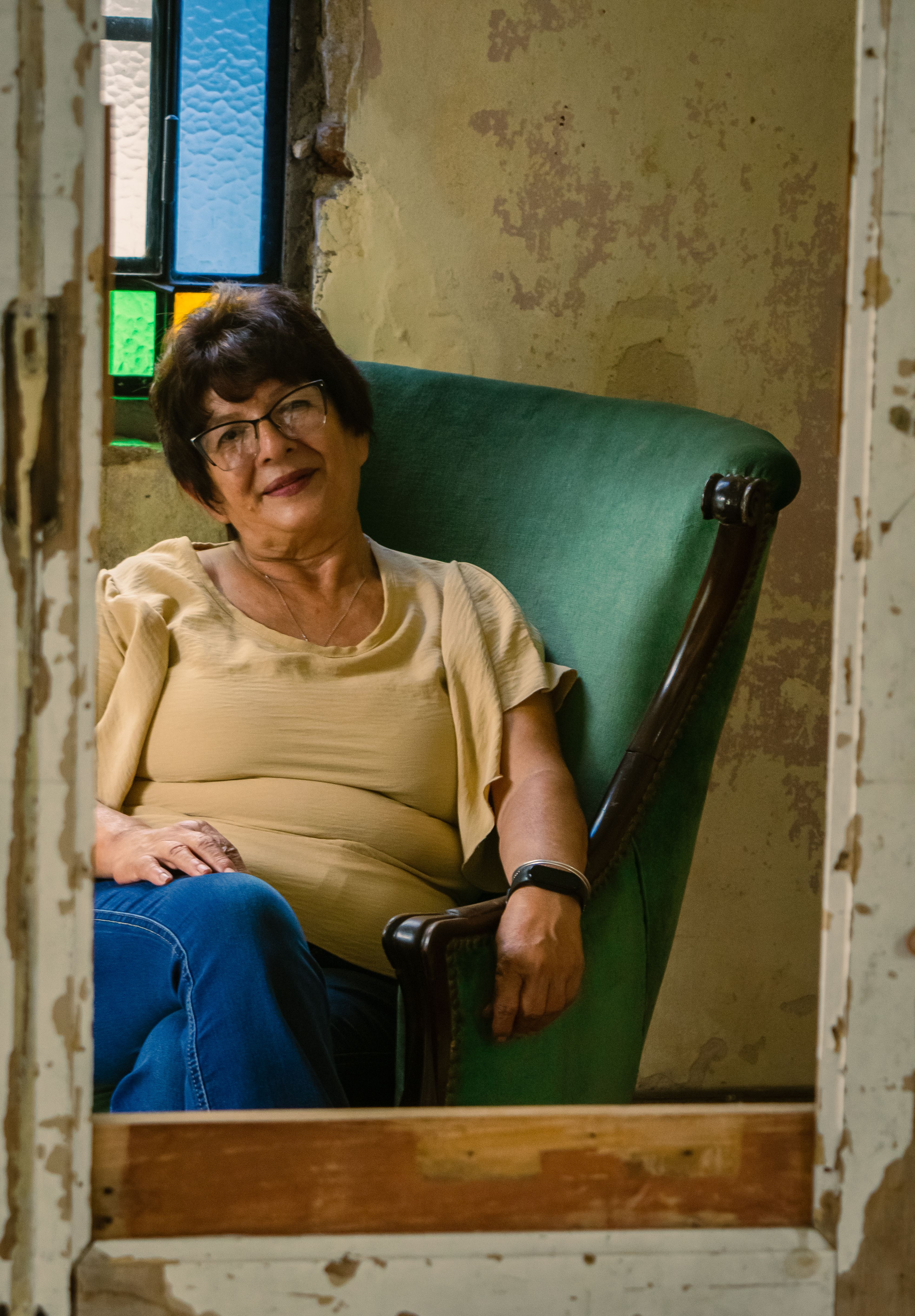 Elderly woman seated in a vintage chair beside stained glass window, indoors.