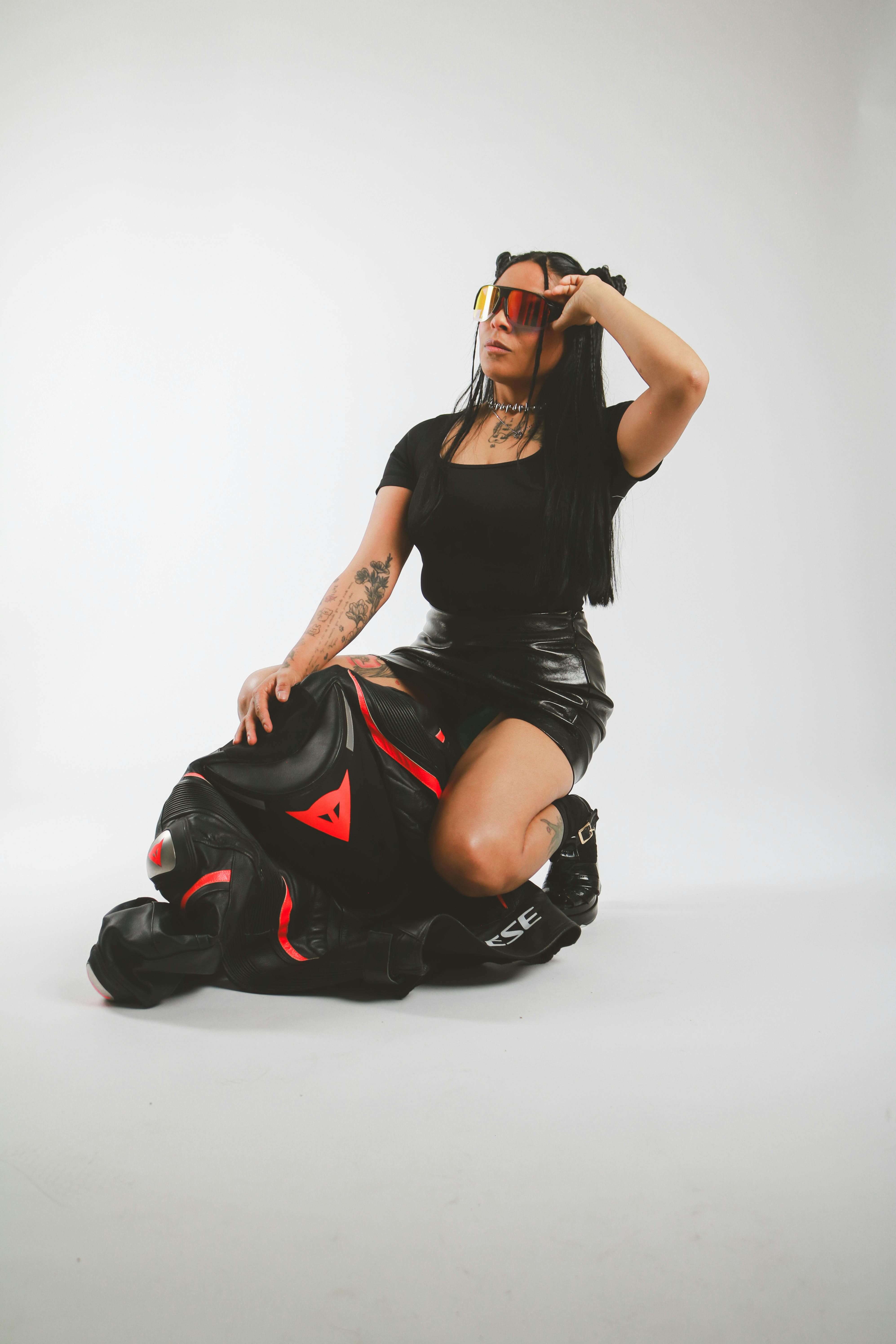 Confident woman posing in leather outfit with motorcycle gear against white background.