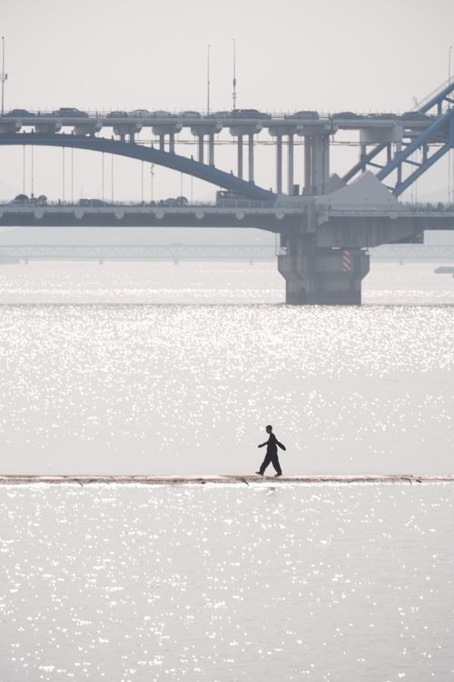 Free A lone figure walks by a shimmering river beneath a large city bridge. Stock Photo