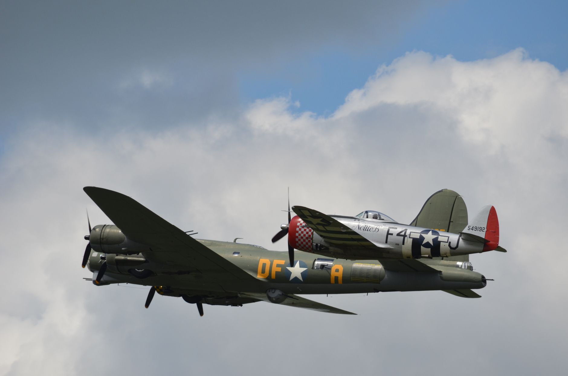 Two vintage military aircrafts, a bomber and a fighter, soaring through a partly cloudy sky, showcasing wartime aviation history.