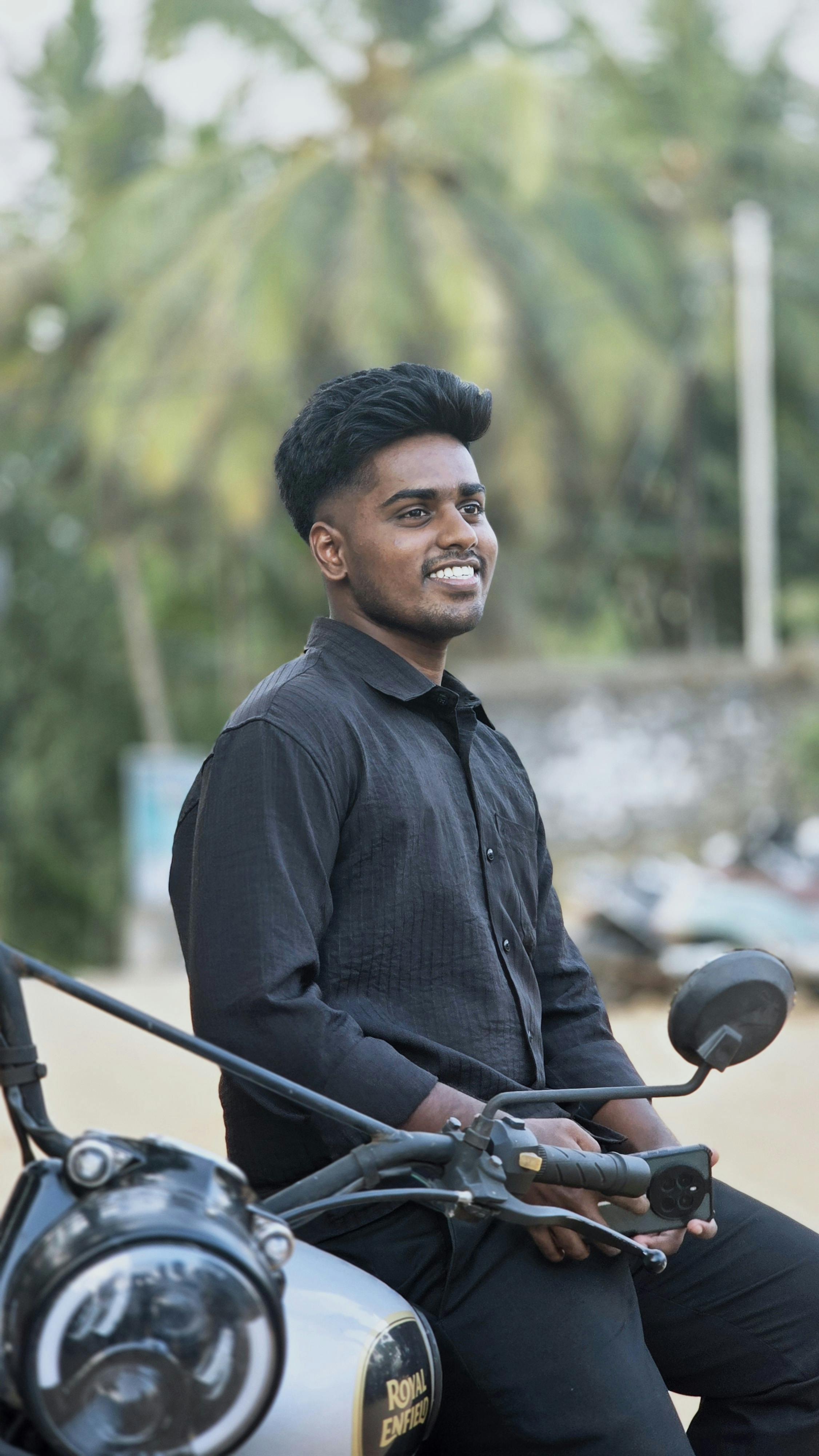 Young man in black shirt sitting on motorcycle with palm trees in background, smiling.
