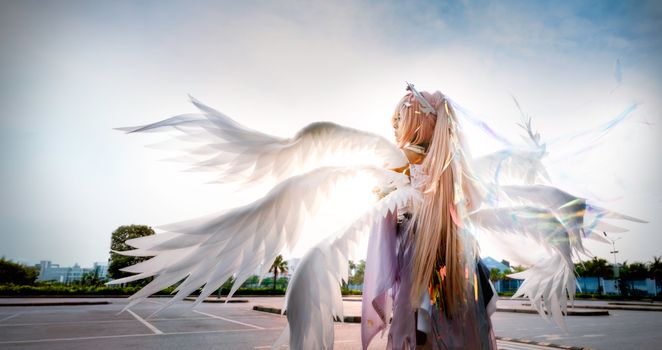 A cosplay character dressed as an angel with large white wings outdoors against a clear sky.