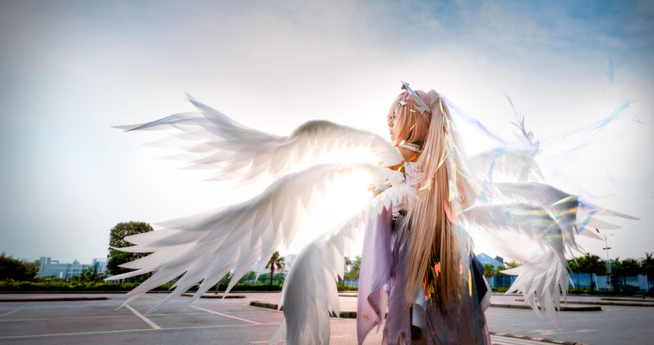 A cosplay character dressed as an angel with large white wings outdoors against a clear sky.
