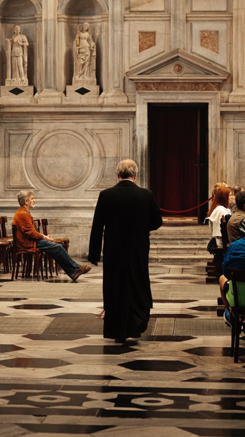 Free A cleric walks through an ornate church interior with people seated, capturing a moment of contemplation. Stock Photo