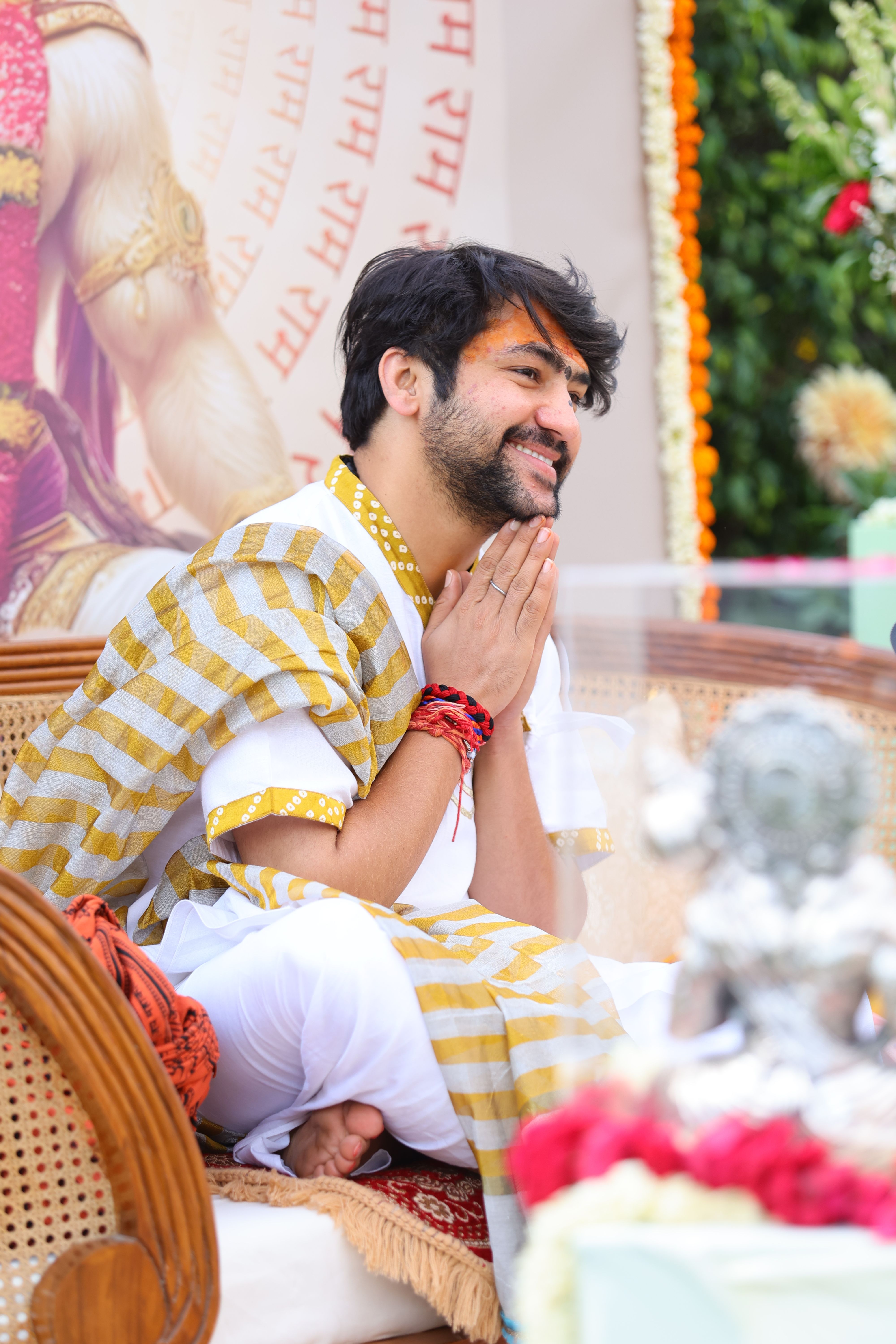 Young man sitting at a colorful outdoor ceremony, wearing traditional attire, hands clasped.