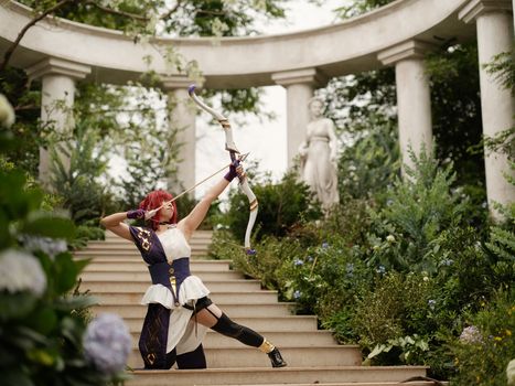 A cosplayer dressed as an archer poses dramatically in a verdant garden with classical architecture.