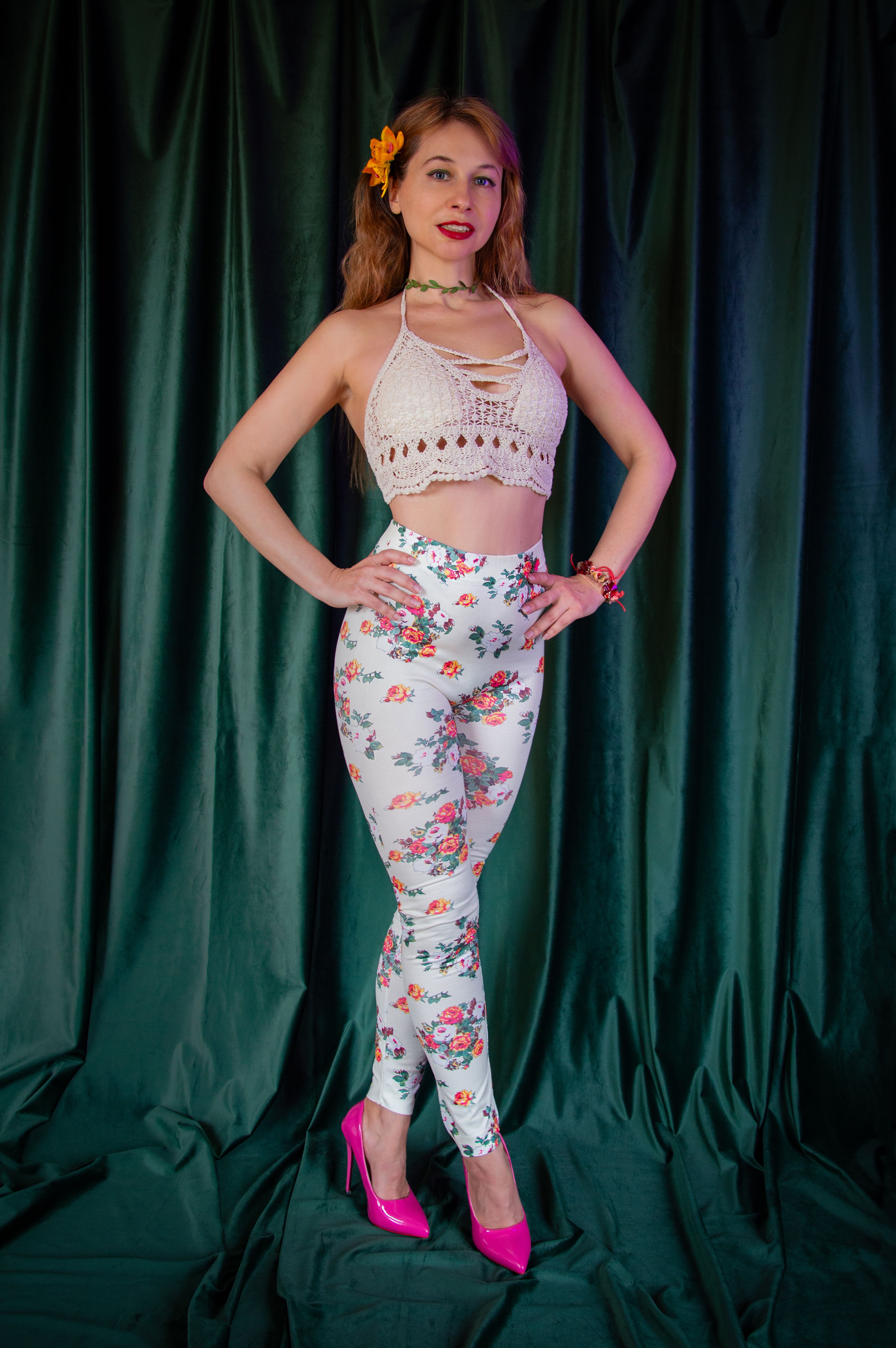 Mulher elegante posando com leggings florais e blusa de crochê contra um fundo verde escuro.