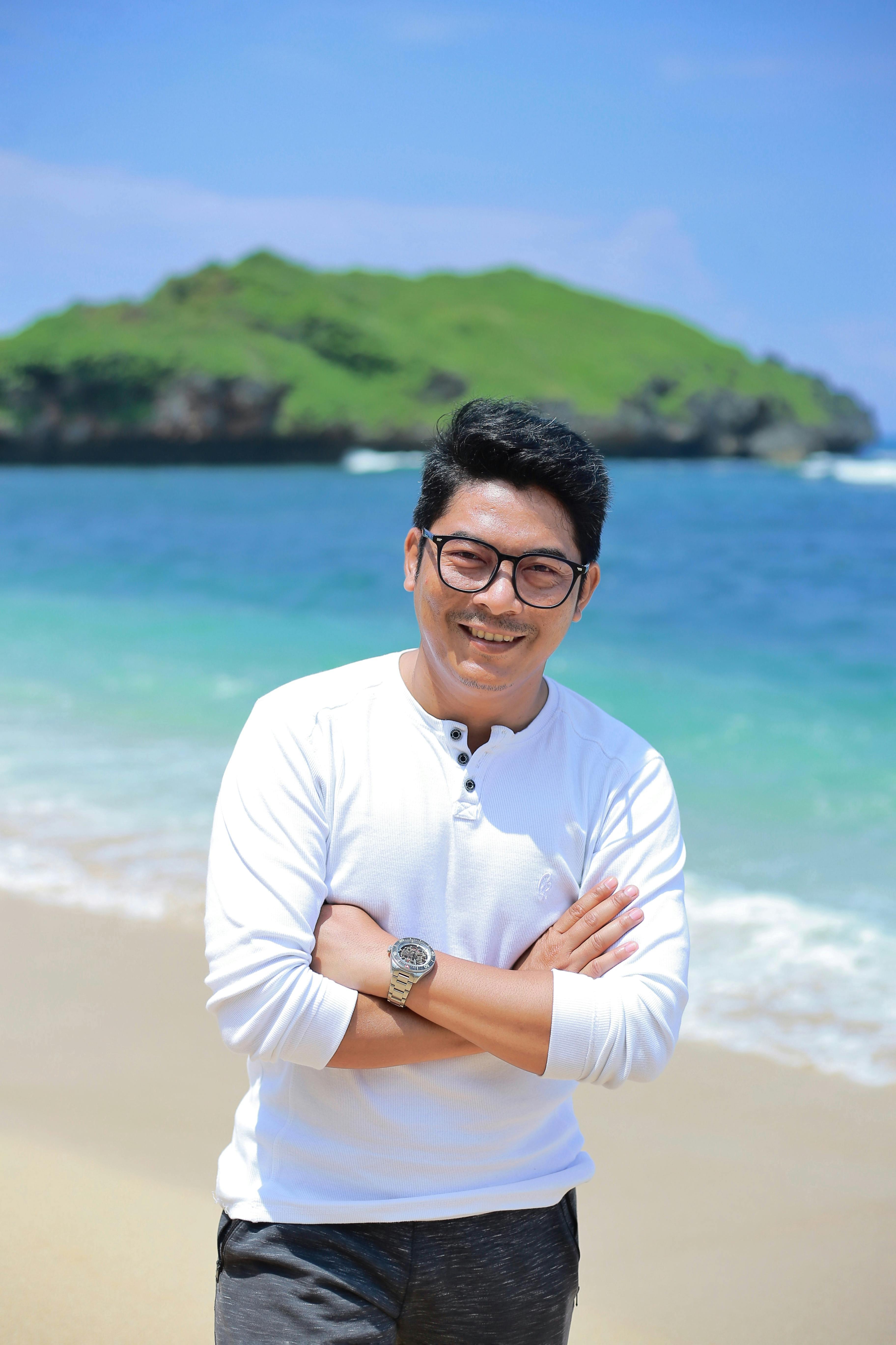 Portrait of a smiling man standing on a sunny beach with blue ocean and green hills.