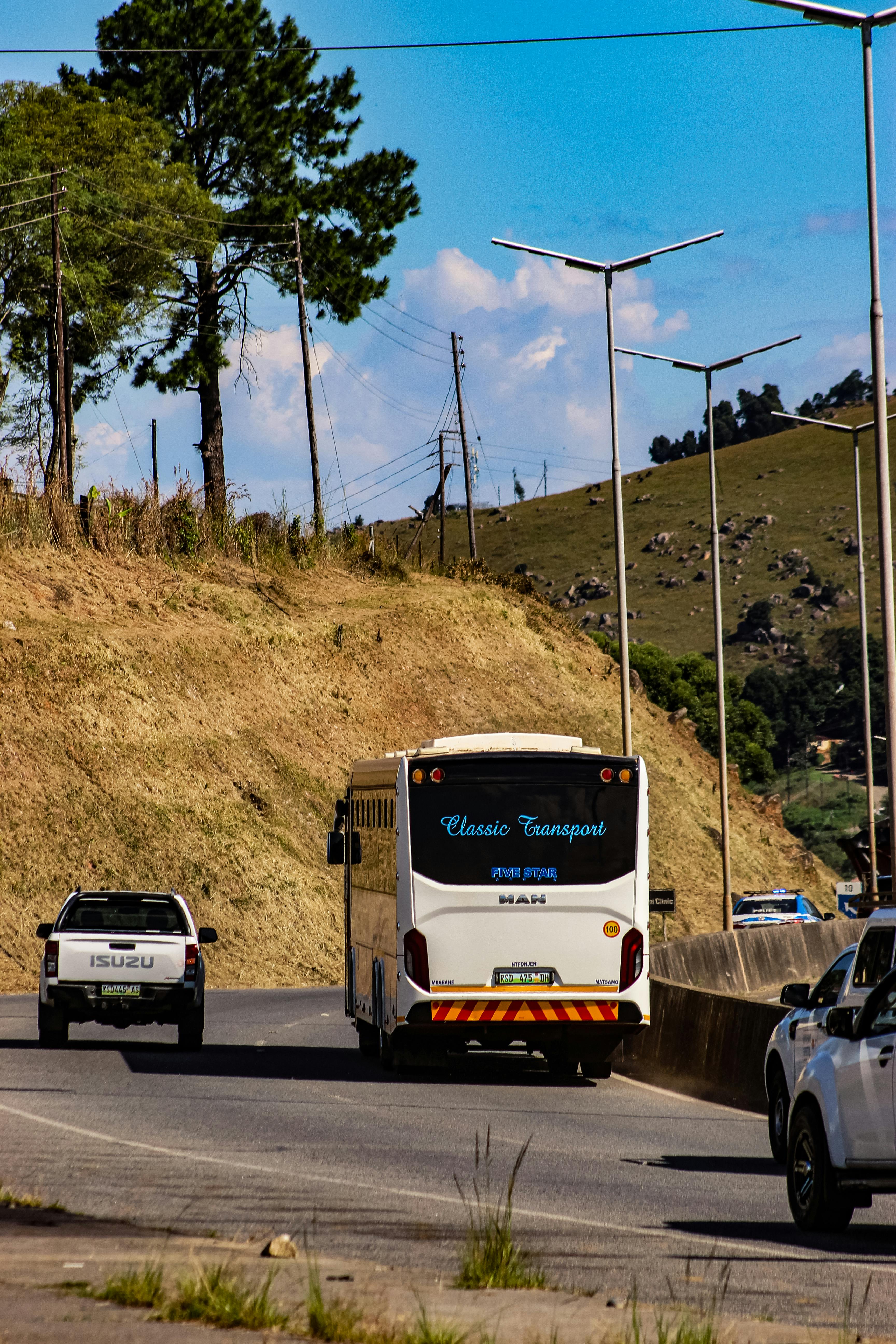 Vehicles travel a curved road in Manzini, Eswatini, surrounded by lush hills and blue skies.
