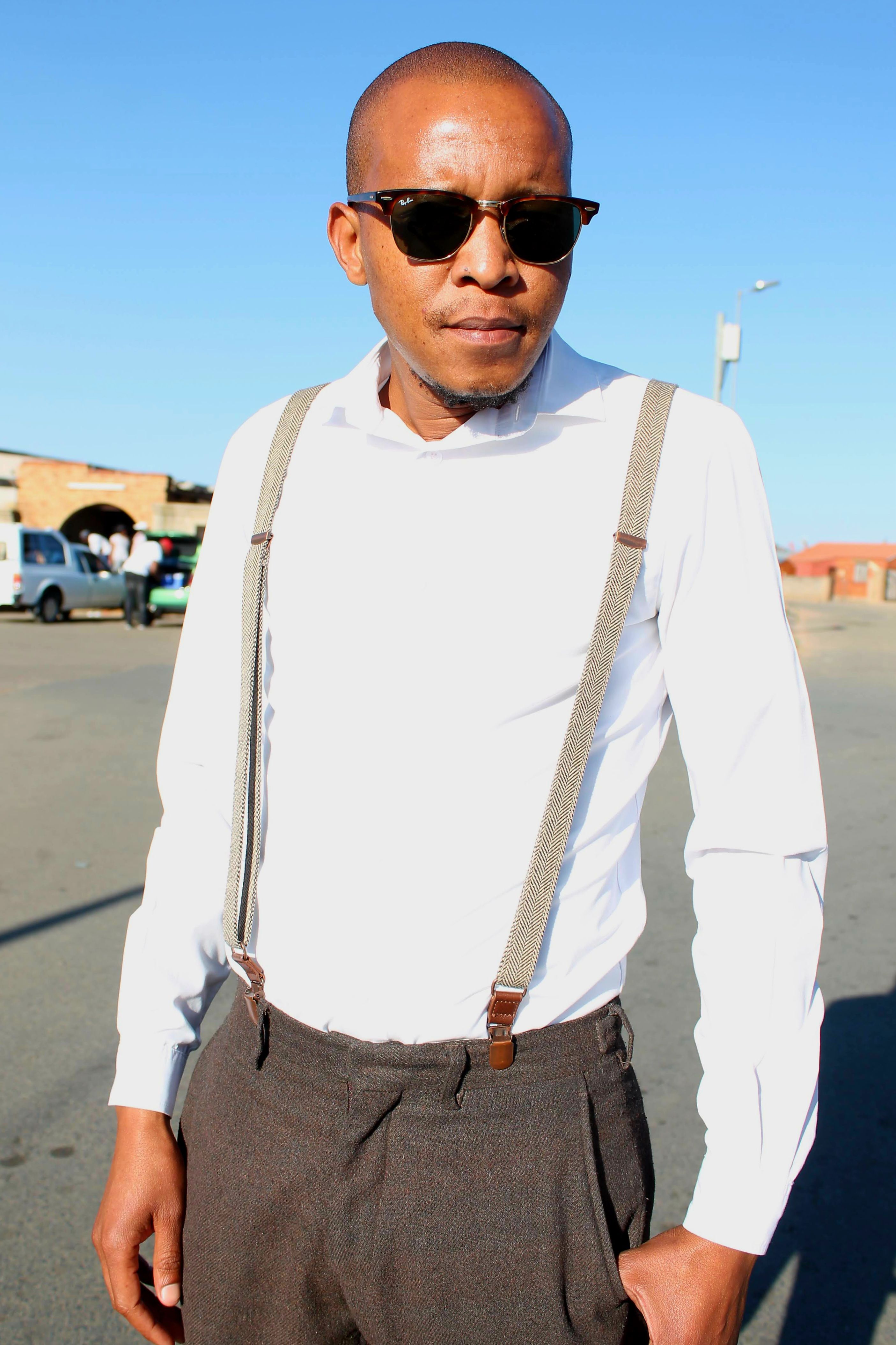 A confident man wearing sunglasses and suspenders stands outdoors, exuding cool style.