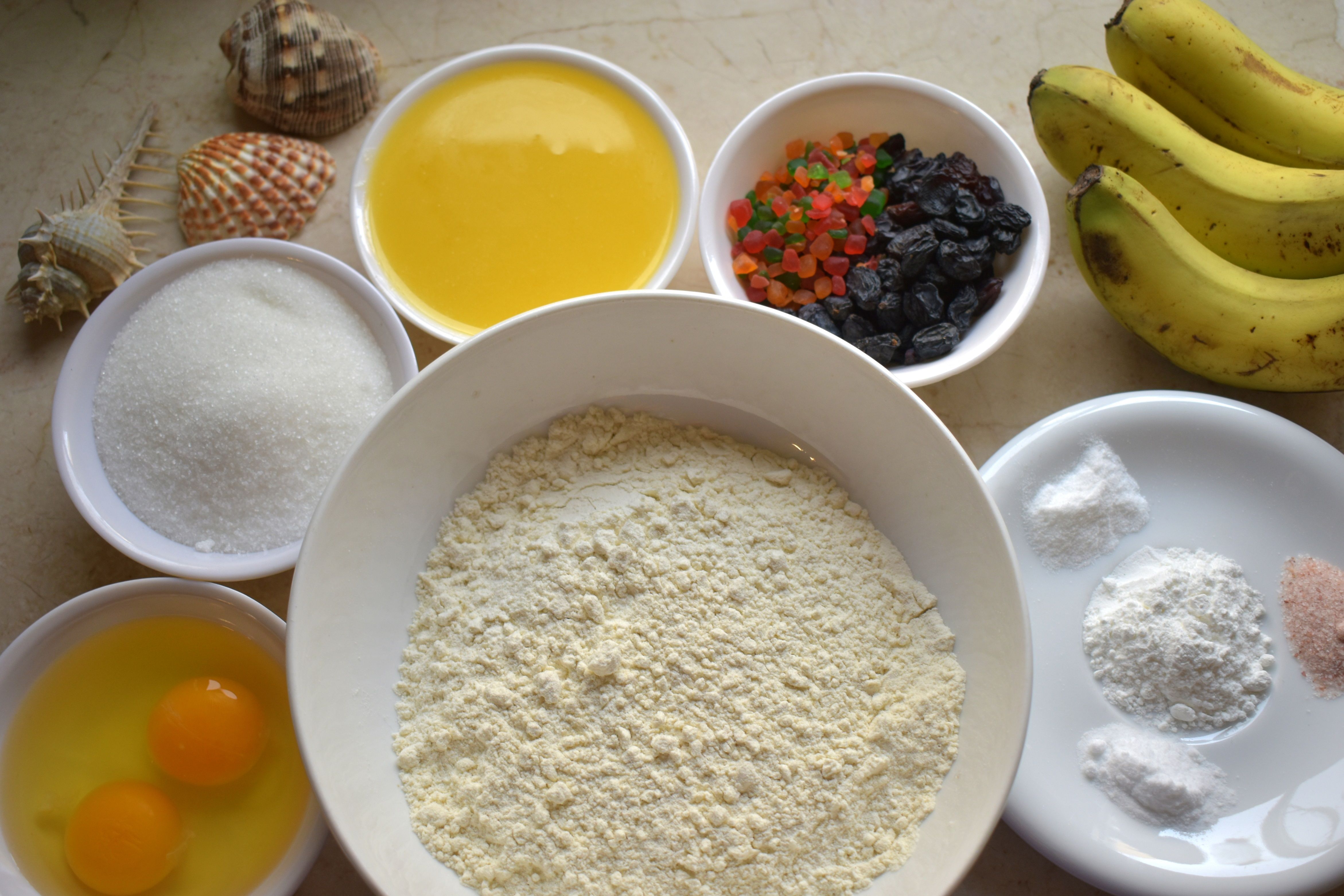 Fresh ingredients for banana bread arranged on marble countertop: ripe bananas, flour, brown sugar, eggs, butter, vanilla, walnuts