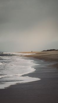 A solitary figure walks along a tranquil beach under a cloudy sky, capturing a sense of peace and solitude.