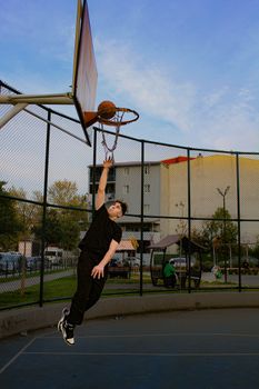 Action shot of a young man performing a slam dunk on an outdoor basketball court during the day.