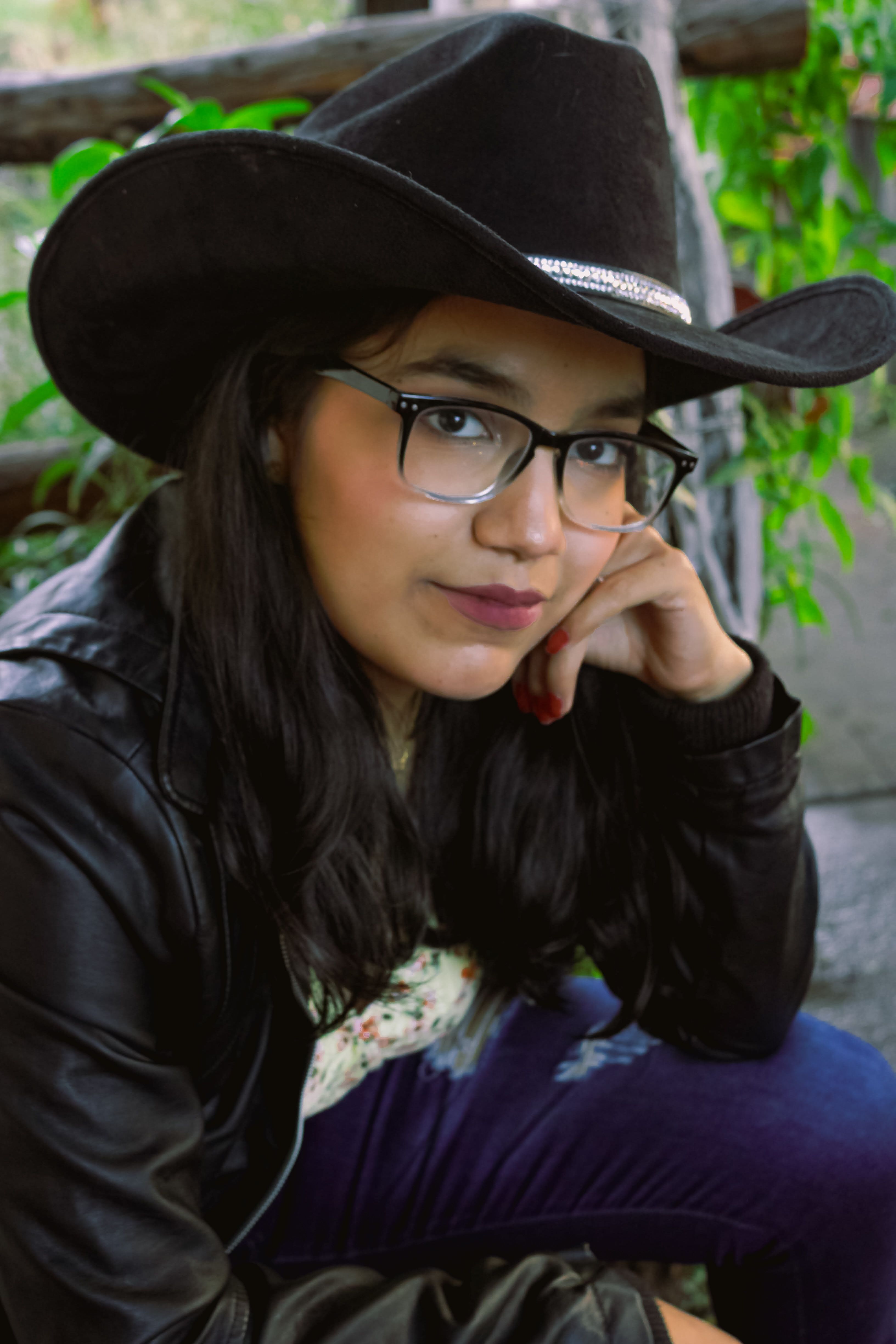 Young woman in glasses wearing a cowboy hat and leather jacket, posing thoughtfully in a relaxed outdoor setting.