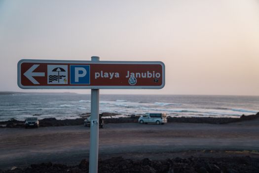 Beach sign at Playa Janubio pointing left under a pastel sky in Lanzarote, Spain with ocean in the background.