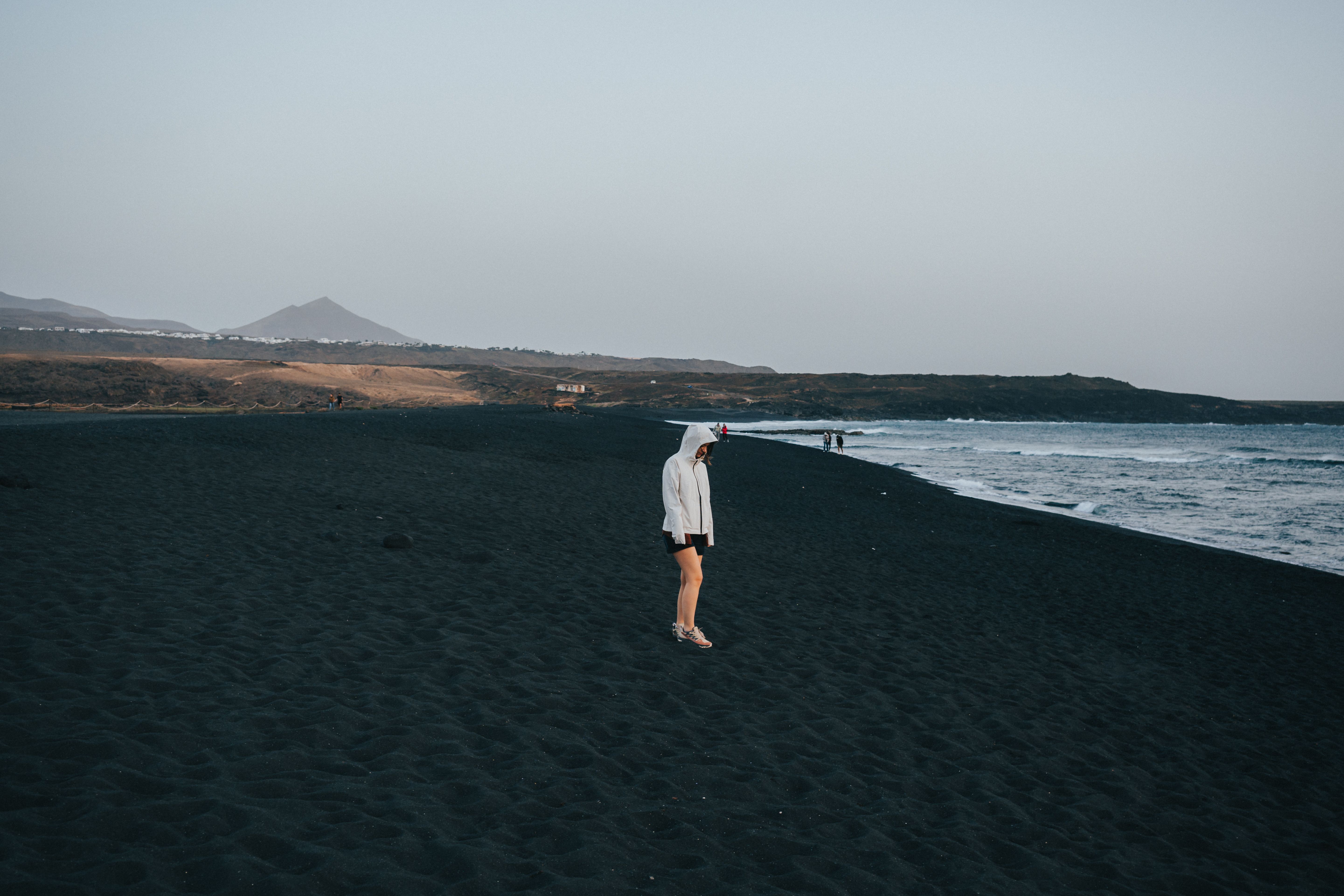 A person stands on the black sand beach of Lanzarote at dusk, highlighting serene coastal solitude.. pexels, person, stands