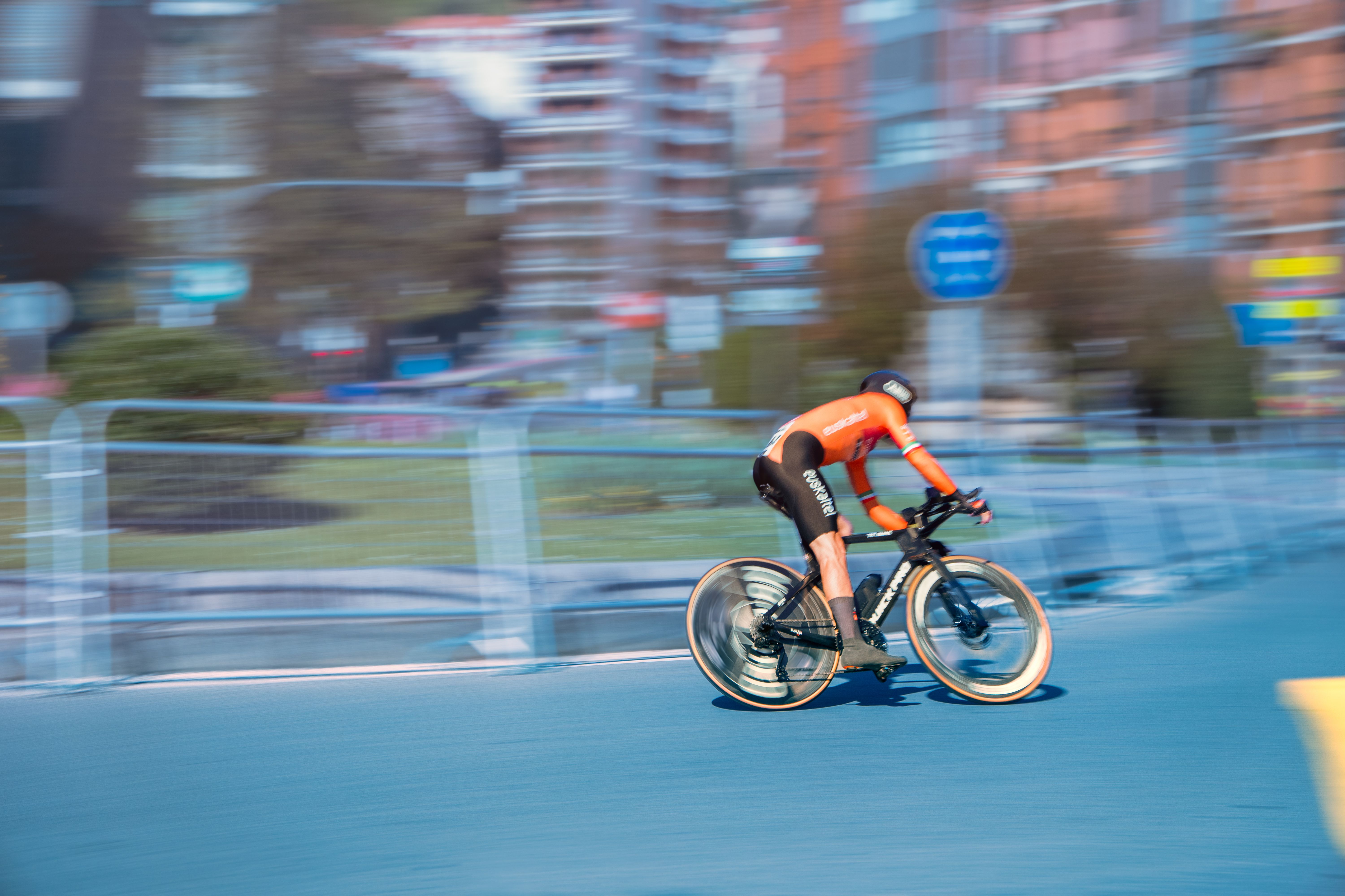 Blurry cyclist races through city streets wearing orange sportswear.