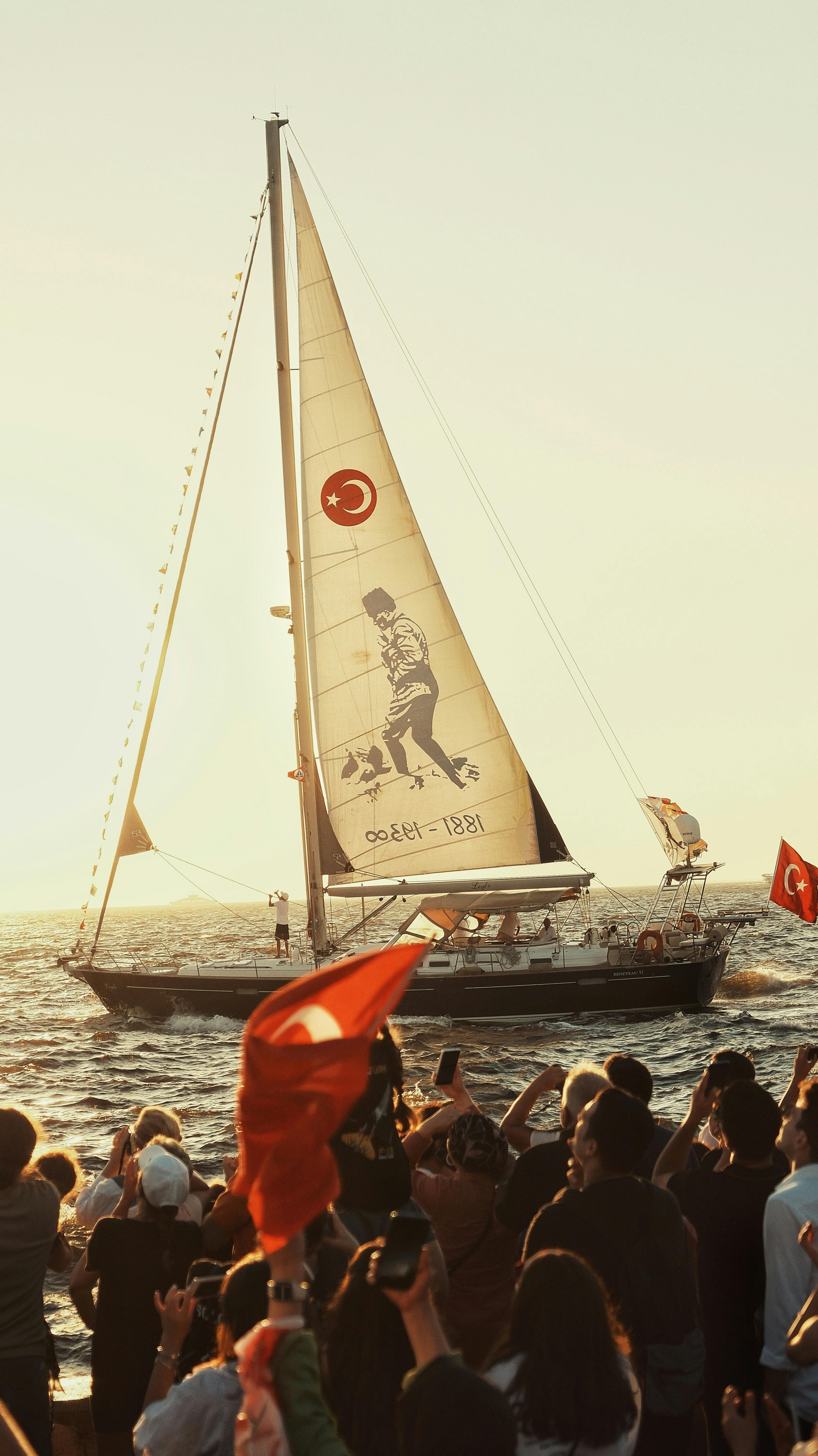 A vibrant sailing event in Izmir, Türkiye, featuring crowd with Turkish flags and a sailboat at sunset.