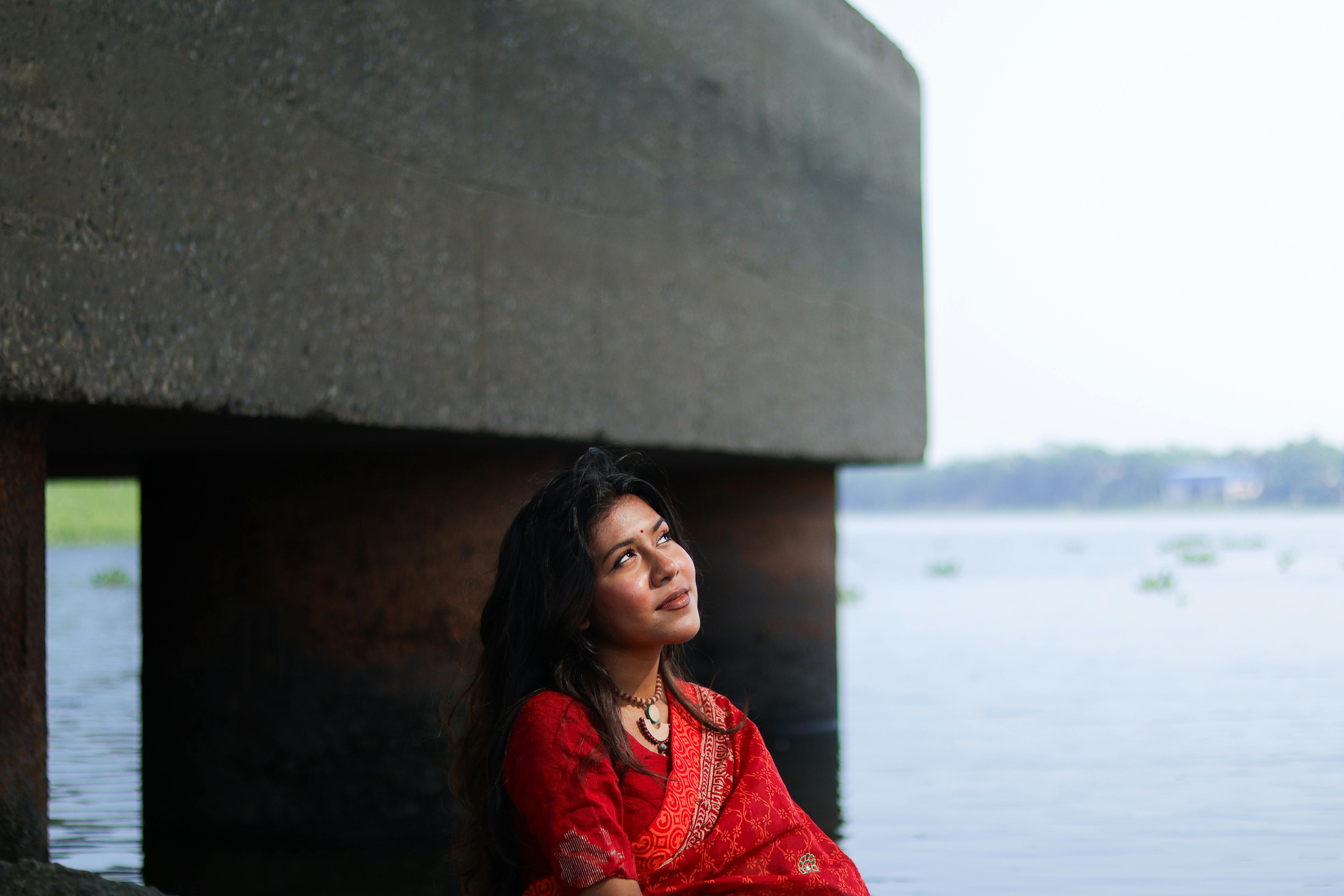 A woman in a red sari gazes thoughtfully by the river under a concrete structure.
