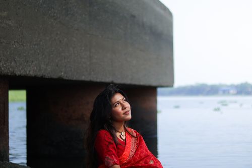 Free A woman in a red sari gazes thoughtfully by the river under a concrete structure. Stock Photo