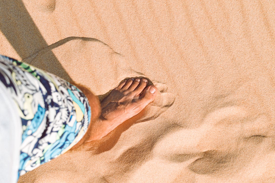 Free Person in Blue and White Floral Shorts Standing on Brown Sand Stock Photo