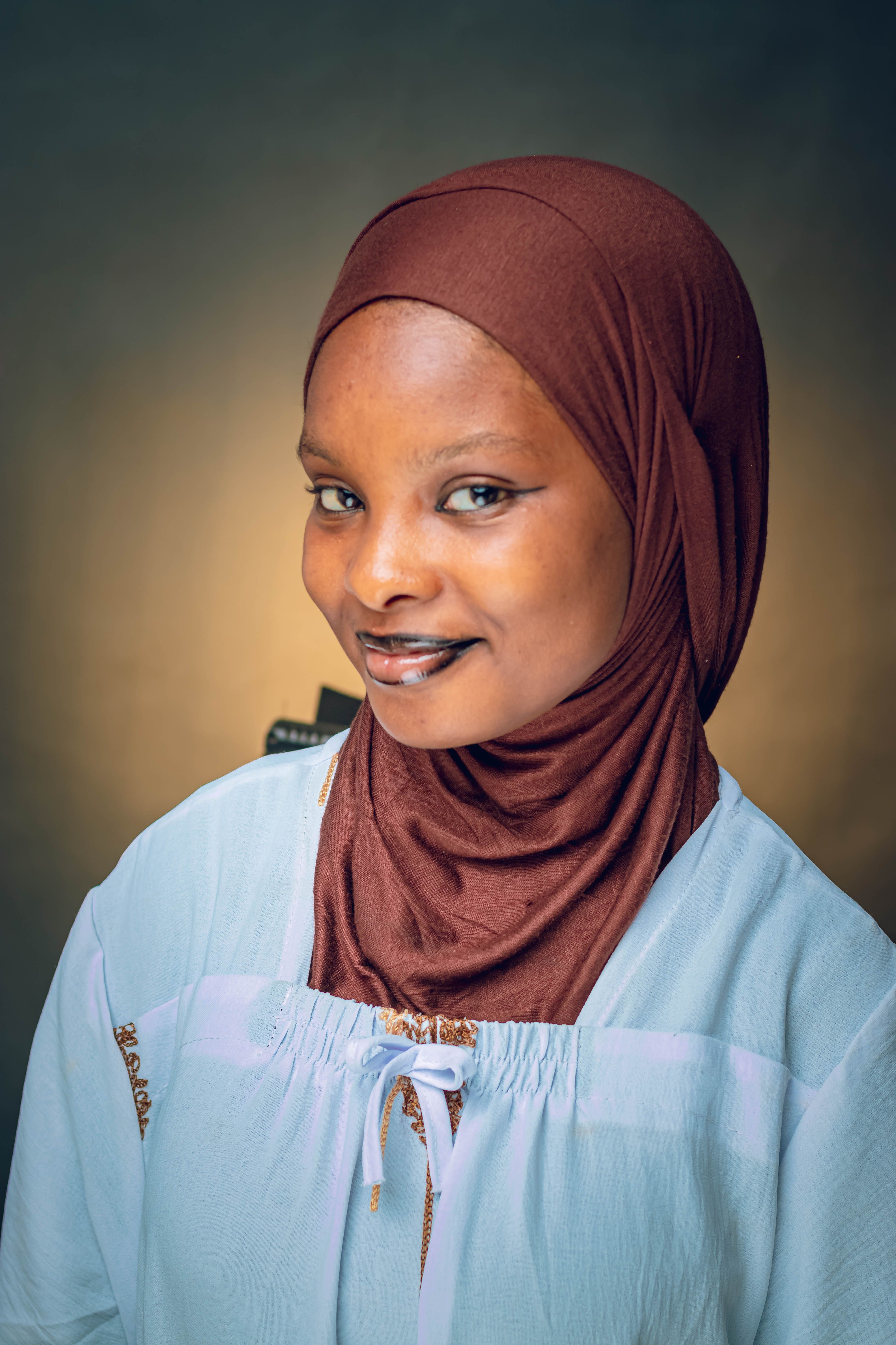 A close-up portrait of a smiling woman wearing a brown hijab and white blouse against a neutral background.