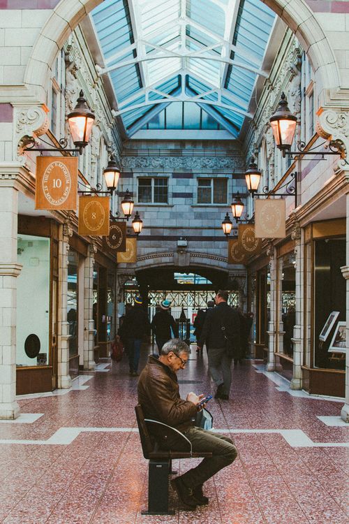 Free An elegant indoor arcade featuring a seated man using a smartphone amidst shoppers, with classic architectural elements. Stock Photo