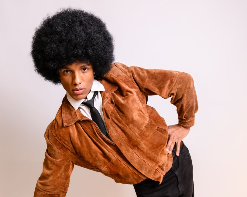 A fashionable man poses with a classic Afro hairstyle, wearing a vintage brown jacket.