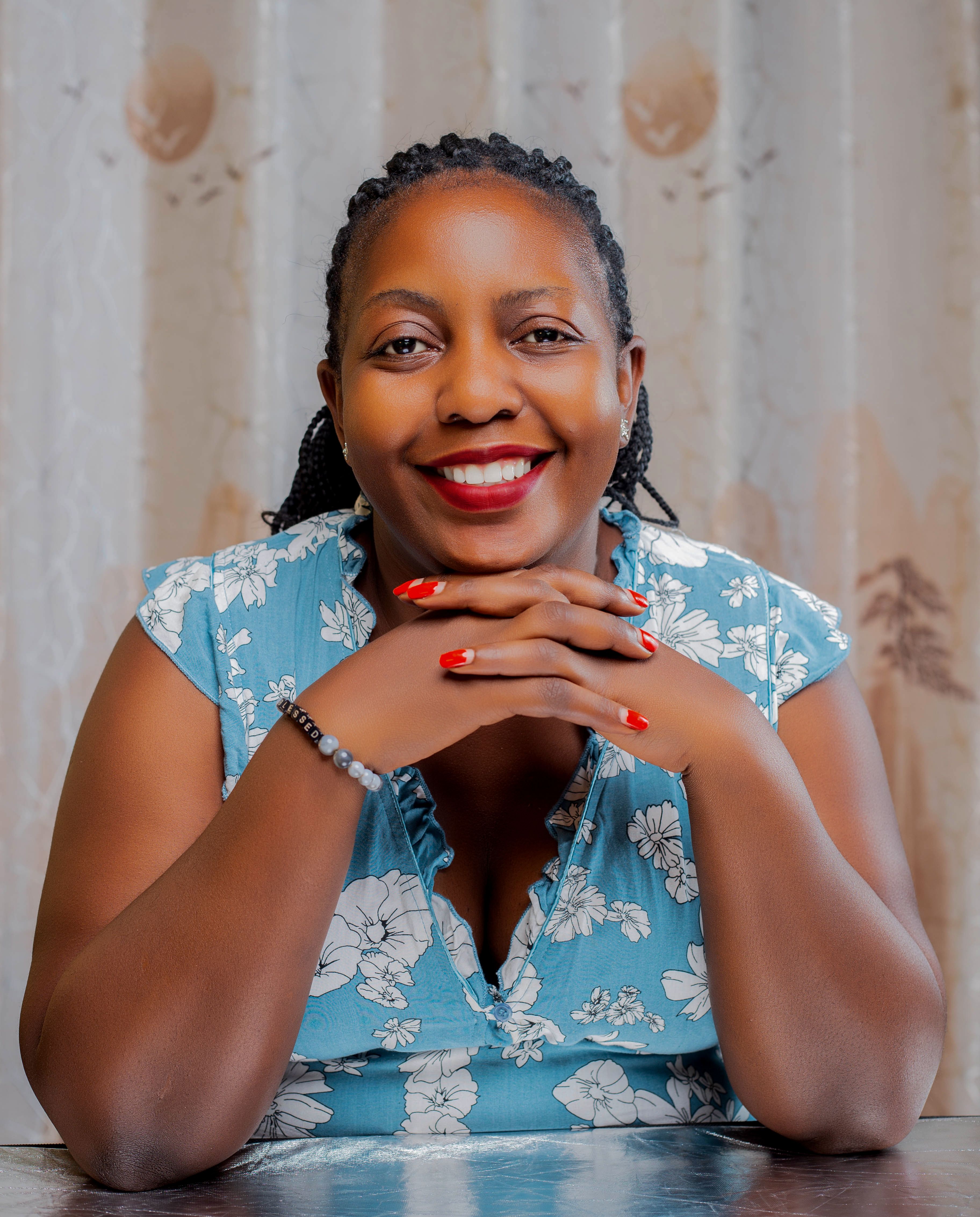 A cheerful woman with red nails sits smiling with hands resting on a table indoors.
