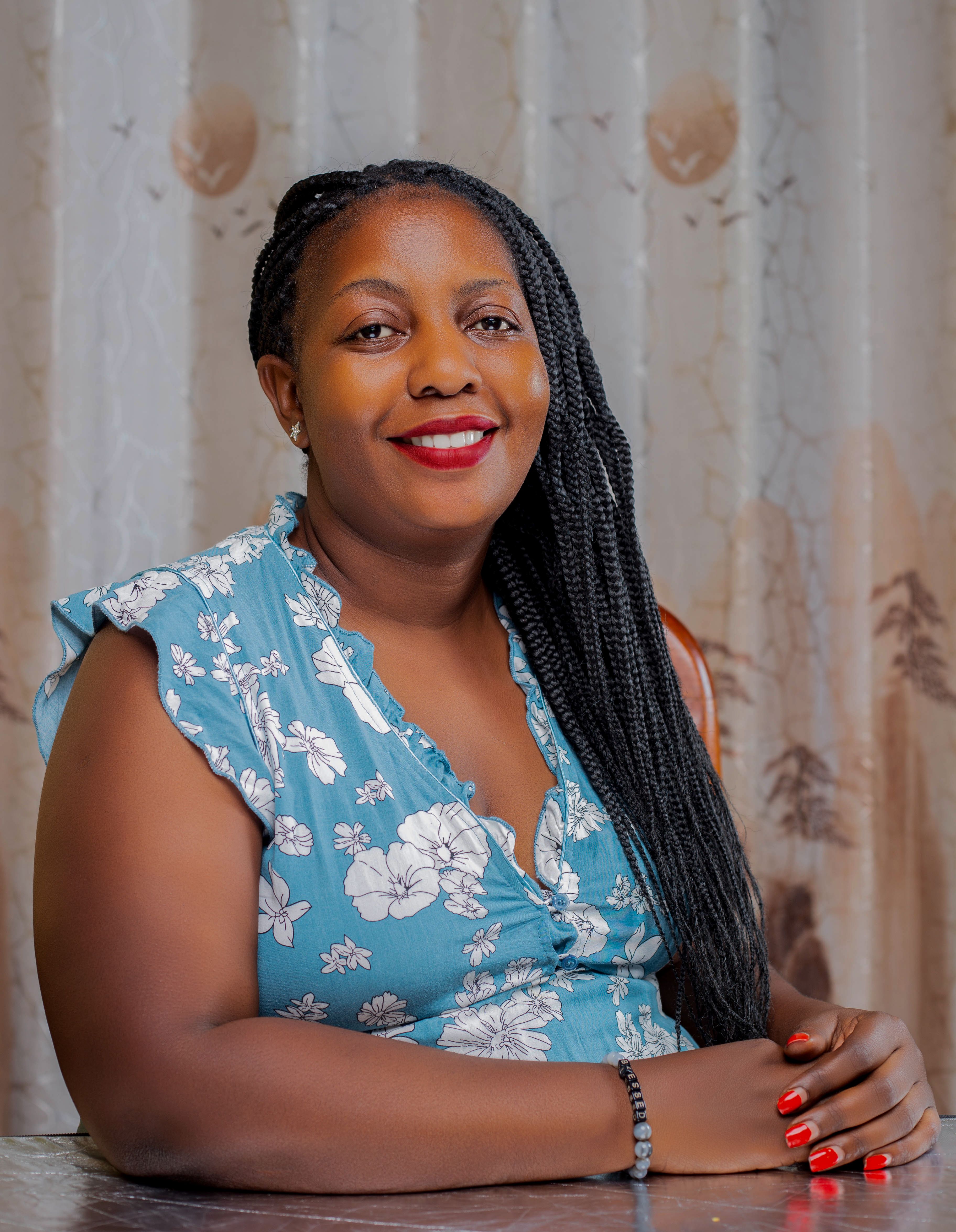 A portrait of a smiling woman in a floral dress with braided hair, sitting indoors.