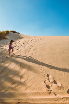 一名女子身着连衣裙，在晴朗的蓝天下攀登沙丘，投下长长的影子