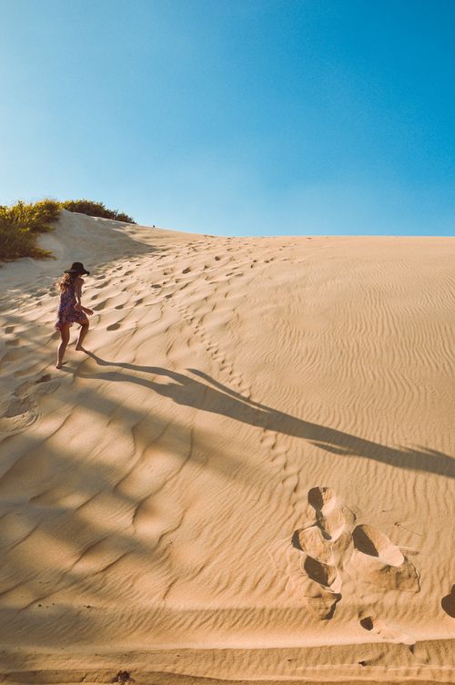 Free A woman in a dress climbs sandy dunes under a clear blue sky, casting long shadows. Stock Photo