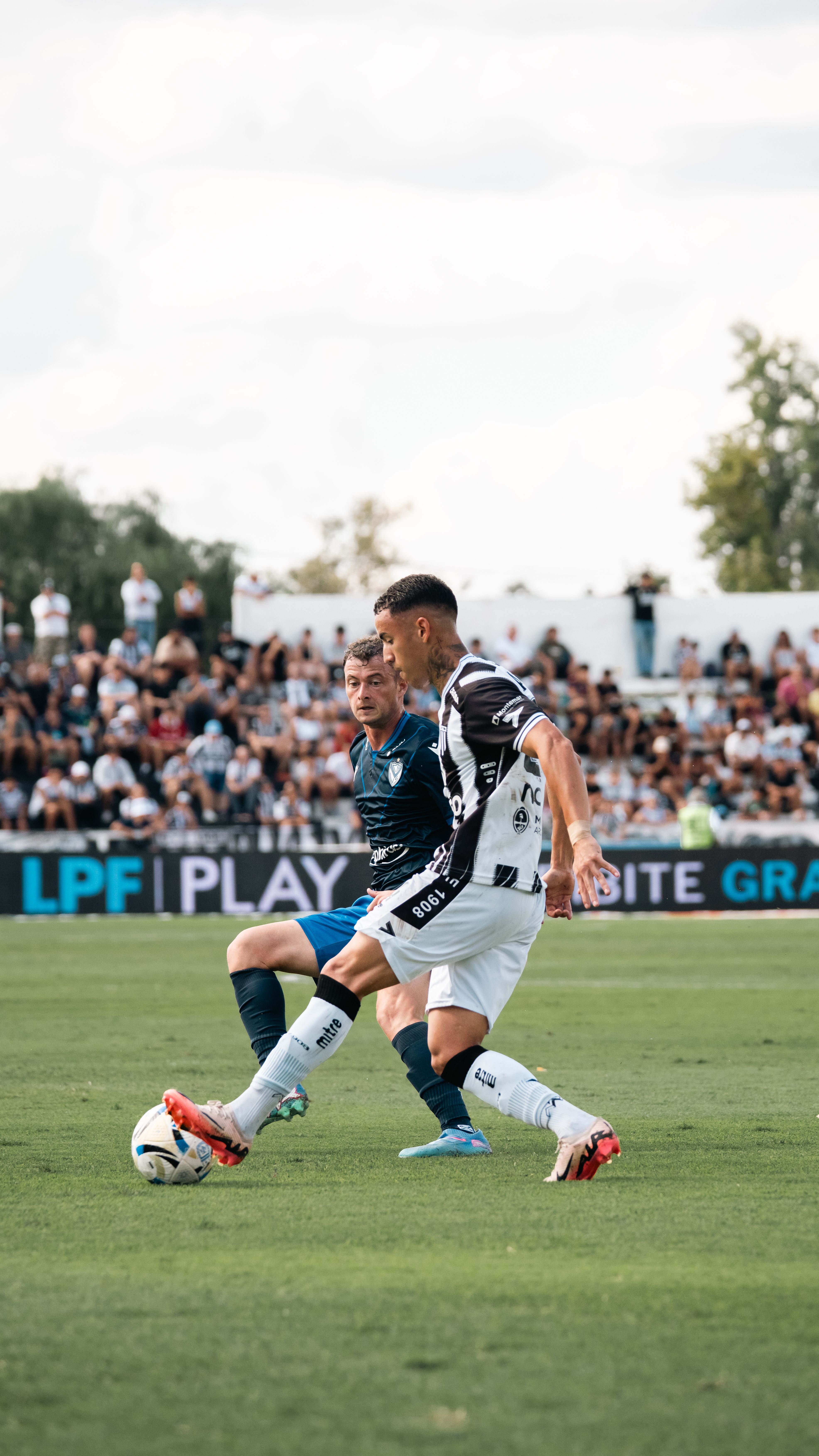 Two soccer players compete fiercely on a bright day with a crowd watching.