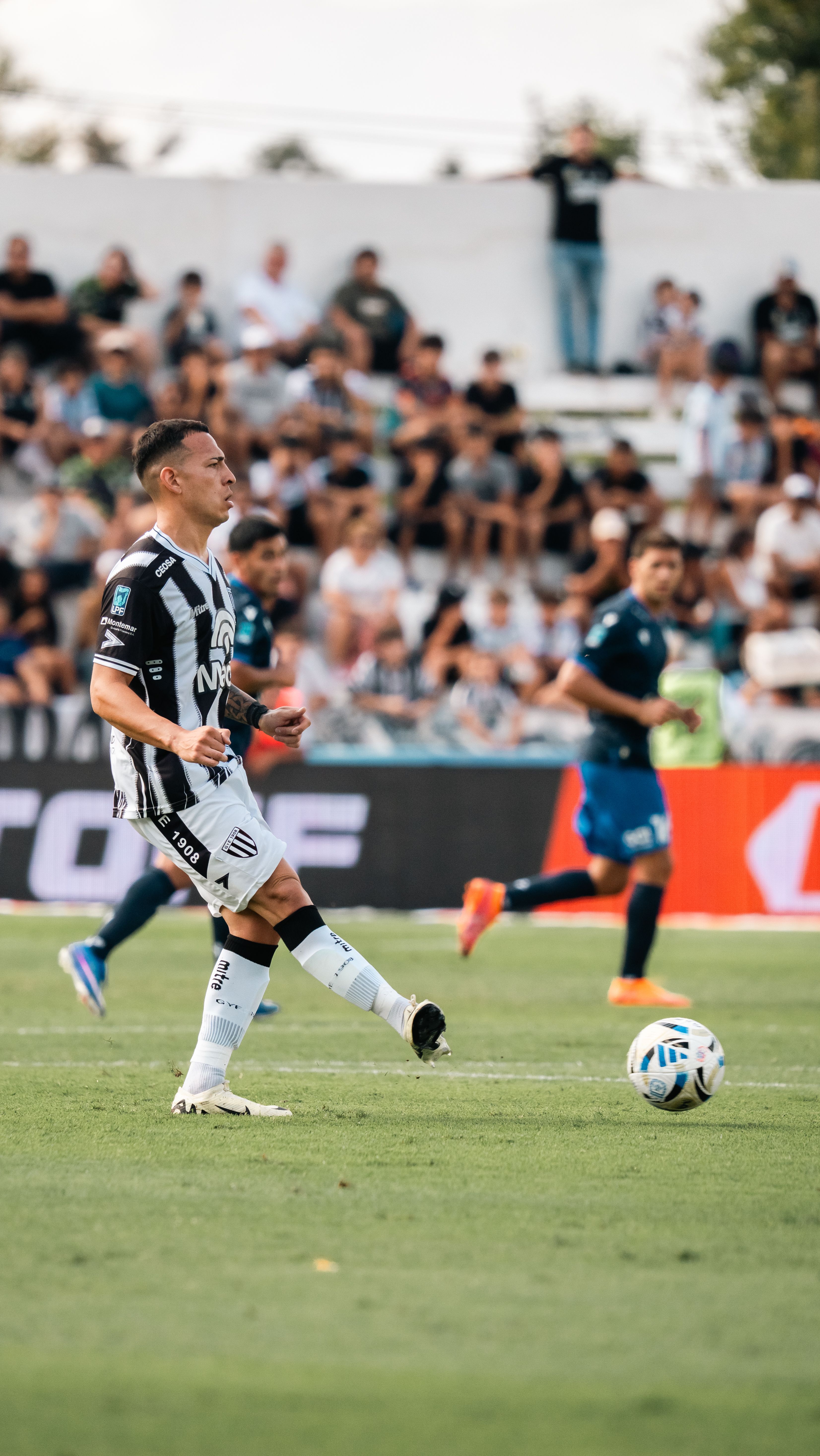 Dynamic scene of players during a competitive soccer match on a sunny day with a crowd in the background.