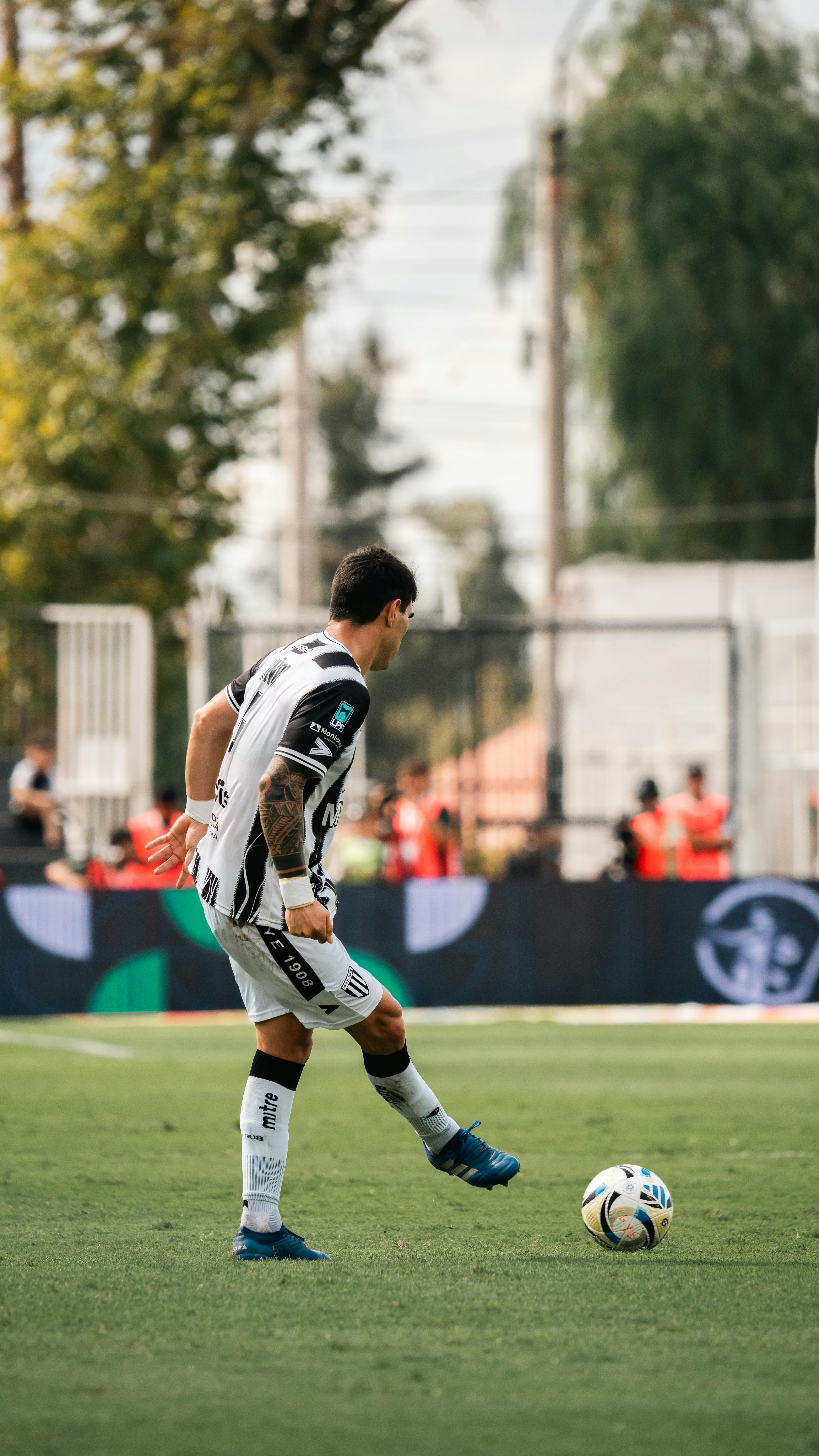 A soccer player in a black and white uniform kicks a ball during a daytime match.