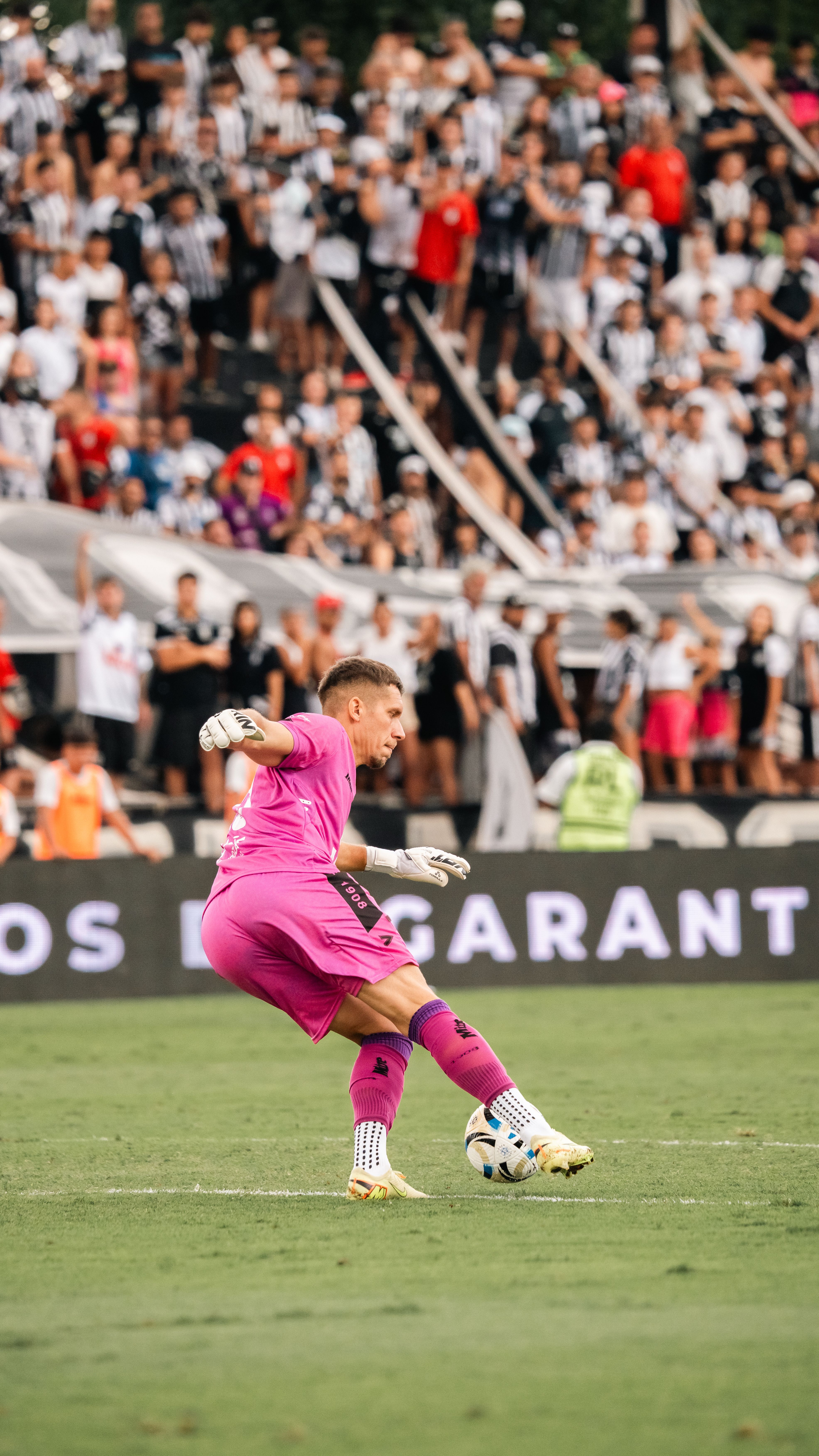 Goalkeeper in pink kit kicking ball during a soccer match in crowded stadium.